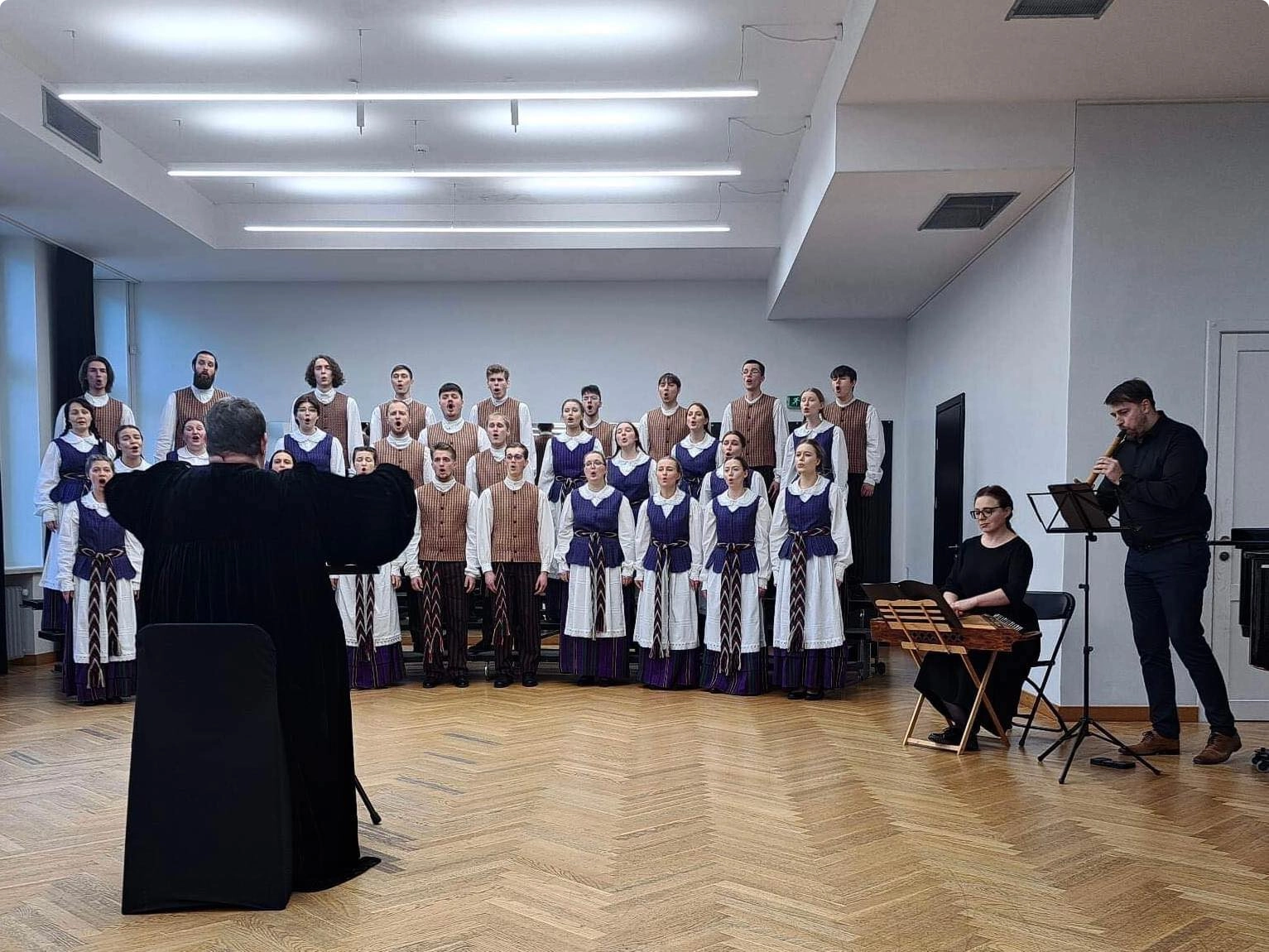 Tautiniais rūbais apsirengę dainuojantys choristai bei jiems diriguojanti maestra, o šalia kanklėmis ir birbyne grojantys pritariamieji muzikantai