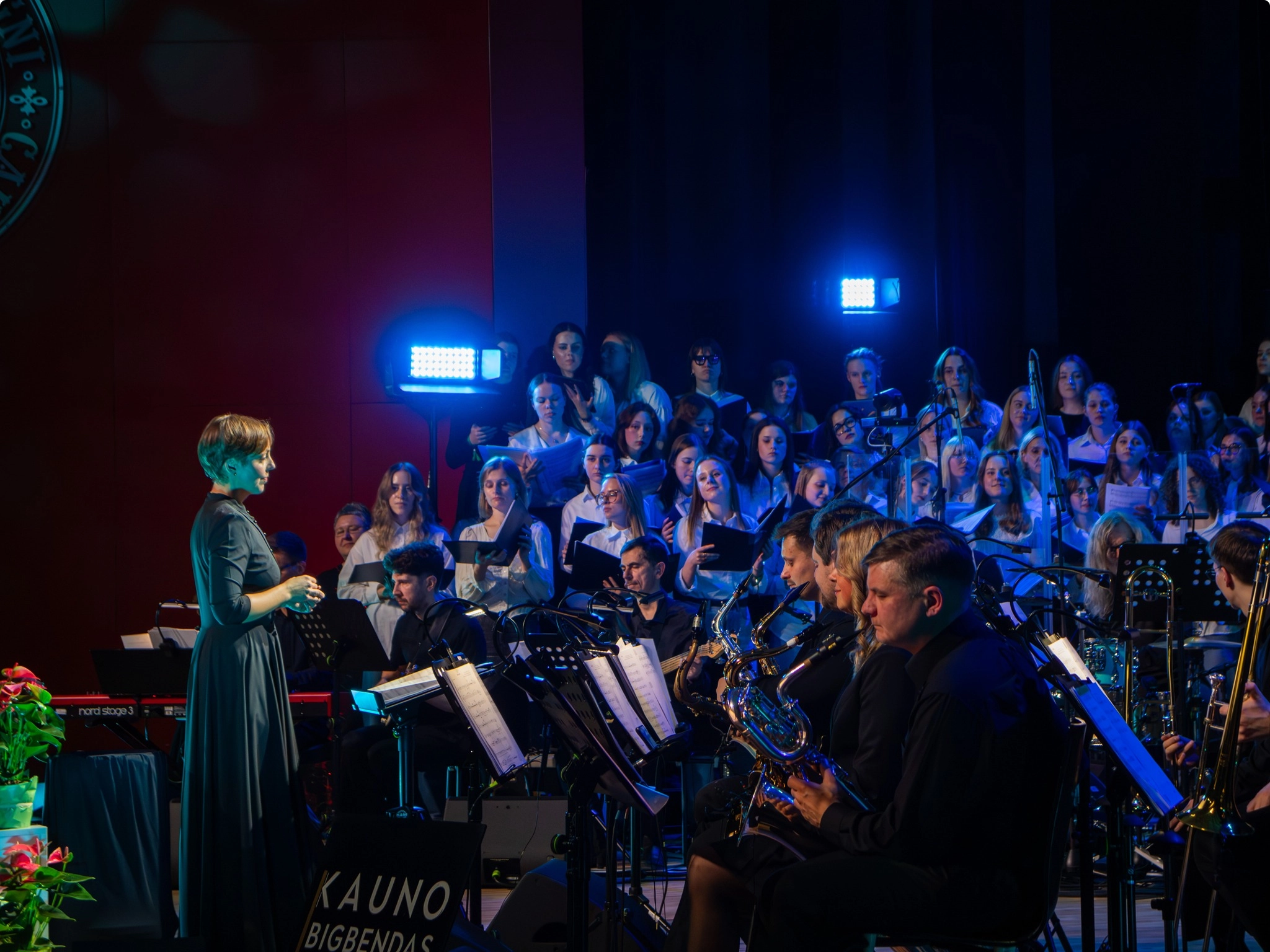 kairėje dirigentė, dešinėje pučiamųjų instrumentų muzikantai, o fone dainininkai studentai