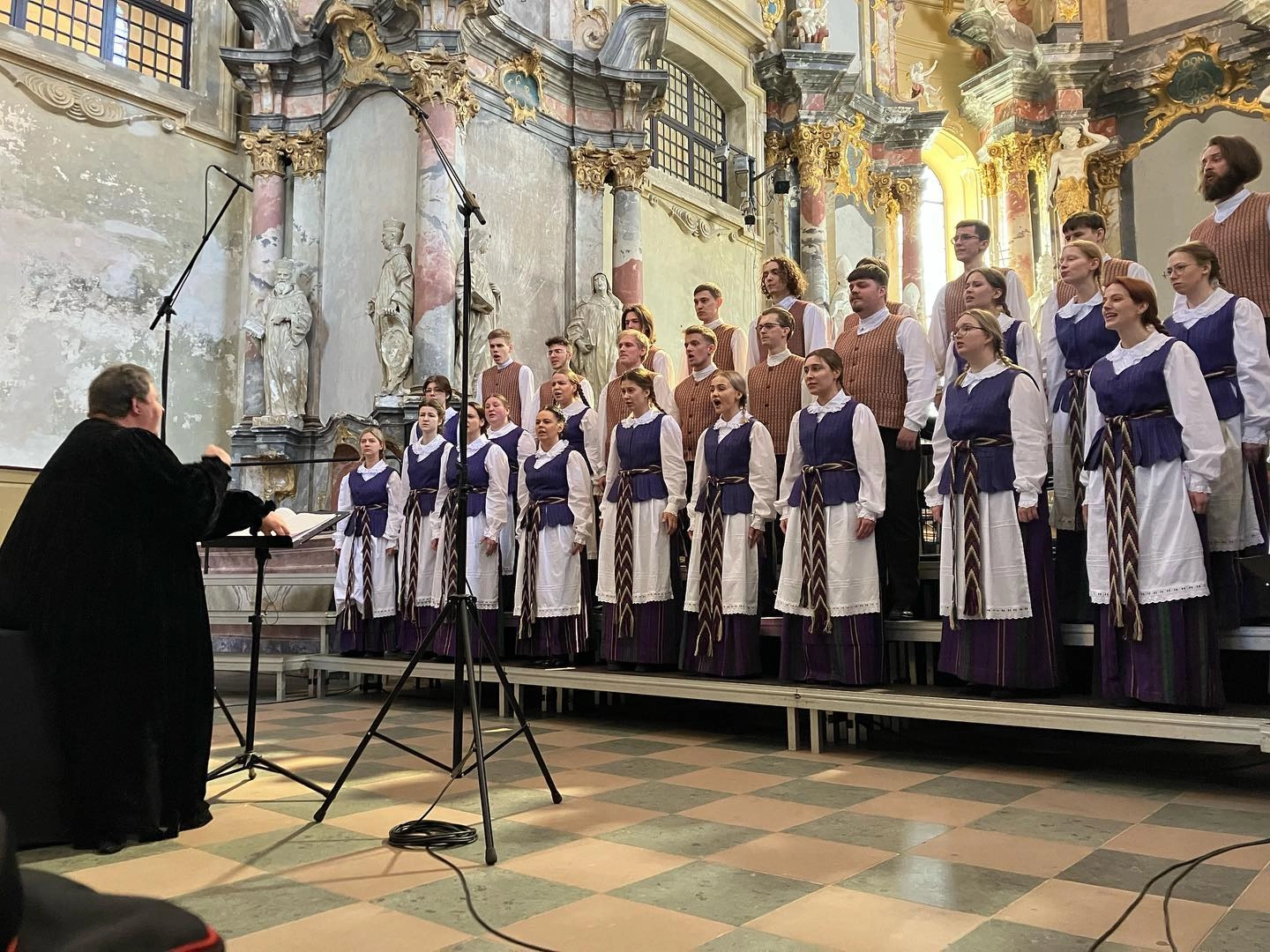 Bažnyčioje tautiniais rūbais apsirengę dainuojantys choristai bei jiems diriguojanti maestra